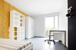  Wohnraum mit immer gleicher Ausstattung aus Bett, Schrank, Regal und Arbeitsplatz. Die Zimmerdecke: weiß gestrichener Sichtbeton  